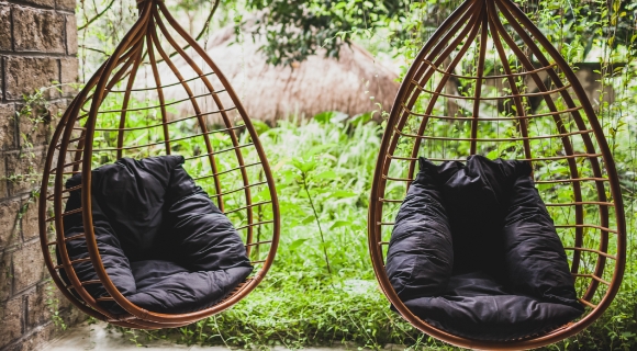 Rattan-H&auml;ngesessel mit Sitzkissen h&auml;ngen nebeneinander auf einer Terrasse im gr&uuml;nen Garten.