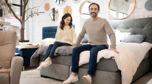 Paar sitzt auf einem Boxspringbett im Einrichtungshaus und testet Sitzhöhe und Komfort – stilvolle Umgebung mit modernen Spiegeln und gemütlicher Atmosphäre