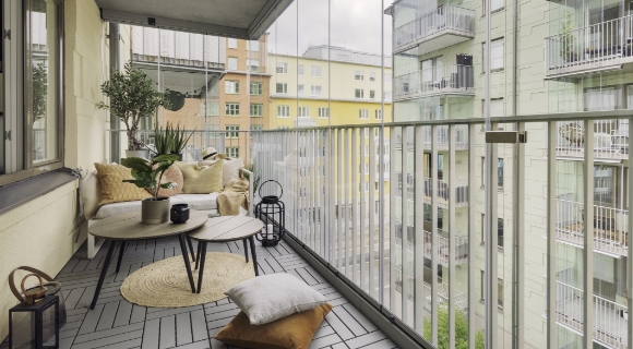 Ein moderner Balkon mit einer Lounge-Ecke, einem runden Tisch und mehreren Kissen. Die Umgebung ist von modernen Geb&auml;uden umgeben.