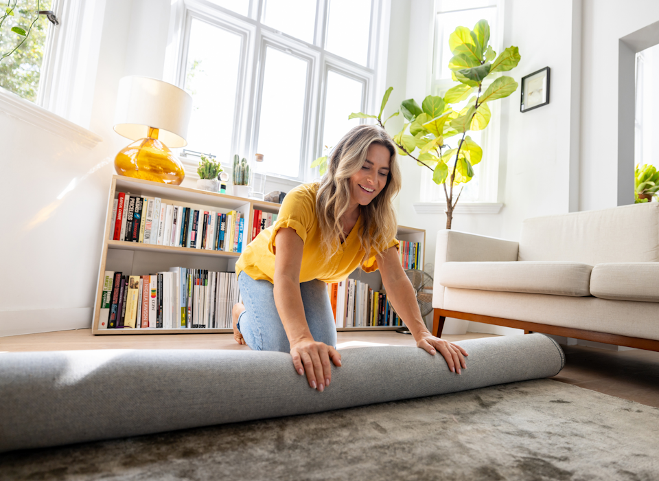 Person rollt Teppich im Wohnzimmer vor Sofa und B&uuml;cherregal aus.