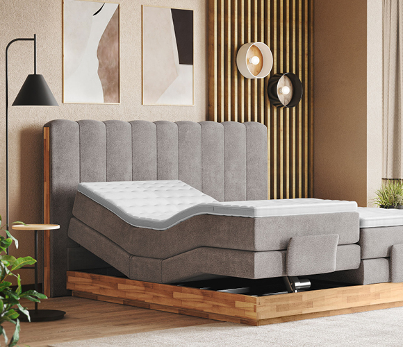 Modernes Boxspringbett in hellem Grau mit hohem, vertikal gestepptem Kopfteil. Die linke Bettseite zeigt eine elektrisch verstellbare Liegefläche: im Rückenbereich angehoben, im Beinbereich leicht angewinkelt, mit weißem, gepolstertem Topper. Das Bett steht auf einem massiven Holzsockel; unter der Matratze ist die Mechanik dezent erkennbar. Links ein schlanker Beistelltisch mit schwarzer Stehleuchte. Im Hintergrund eine Akzentwand aus vertikalen Holzlamellen, runde Wand­leuchten und abstrakte Bilder. Warme Naturtöne, heller Boden und etwas Grün sorgen für ein gemütlich-modernes Ambiente