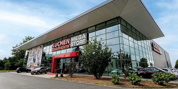Das Bild zeigt die Au&szlig;enansicht eines modernen K&uuml;chenfachmarkts mit gro&szlig;fl&auml;chiger Glasfassade und dem Schriftzug &bdquo;K&uuml;chen Schaffrath Mega Store&ldquo; &uuml;ber dem Eingangsbereich. Der Eingang ist durch einen farblich abgesetzten Rahmen hervorgehoben. Vor dem Geb&auml;ude befinden sich bepflanzte Gr&uuml;nfl&auml;chen sowie mehrere parkende Fahrzeuge. Die Aufnahme zeigt das Geb&auml;ude bei Tageslicht aus seitlicher Perspektive.