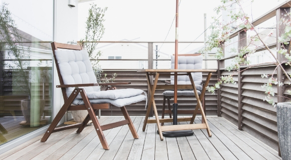 Holzbalkon mit Tisch, zwei gepolsterten St&uuml;hlen und Sonnenschirm neben Pflanzk&uuml;beln und Sichtschutz aus Holz
