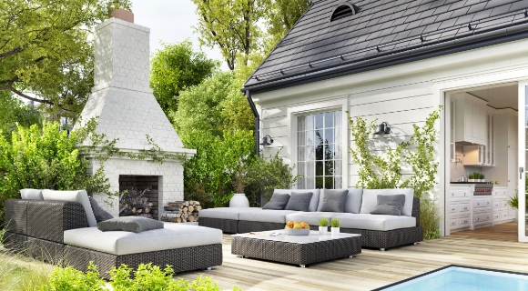 Große Terrassensitzgruppe mit Polstern in Grau neben einem weißen Kaminofen und einem offenen Zugang zur Küche.