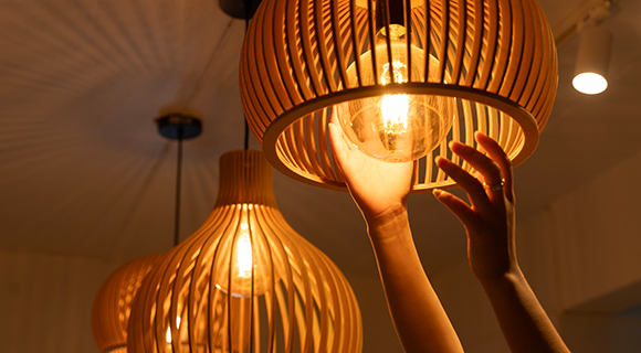 Drei eingeschaltete Deckenlampen mit Holzlamellen geben warmes Licht, eine Hand richtet eine Glühbirne in einer der Leuchten aus.