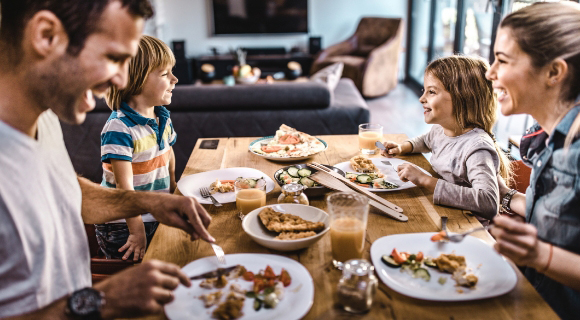 Junge Familie sitzt lachend am Esstisch im Wohnzimmer und isst gemeinsam. Zwei Erwachsene und zwei Kinder genießen verschiedene Speisen und Getränke in heller, wohnlicher Atmosphäre.