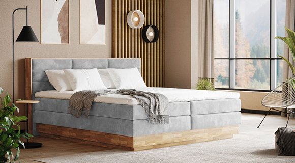 Modernes Schlafzimmer mit großem Boxspringbett in hellem Grau und gepolstertem, segmentiertem Kopfteil. Das Bett steht auf einem massiven Holzsockel in warmen Naturtönen; weiße Bettwäsche und eine locker über die Kante gelegte graue Strickdecke sorgen für Gemütlichkeit. Links ein schlanker Beistelltisch mit schwarzer Leselampe, dahinter eine Akzentwand aus vertikalen Holzlamellen mit runder Wanddeko/Leuchte; daneben abstrakte Bilder in Beige- und Sandtönen. Rechts öffnet eine bodentiefe Fensterfront den Blick auf Bäume, der Raum hat hellen Teppichboden und dezente Pflanzen. Der Gesamteindruck ist warm, modern und naturverbunden