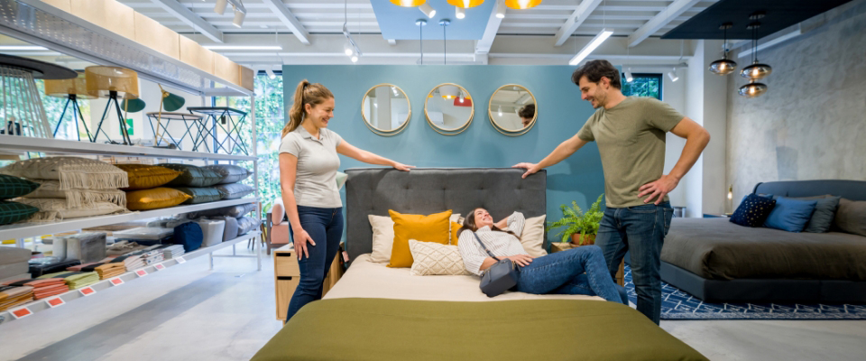 Einrichtungsexpertin berät ein junges Paar beim Probeliegen auf einem Polsterbett in einer stilvoll dekorierten Bettenausstellung mit Wohnaccessoires