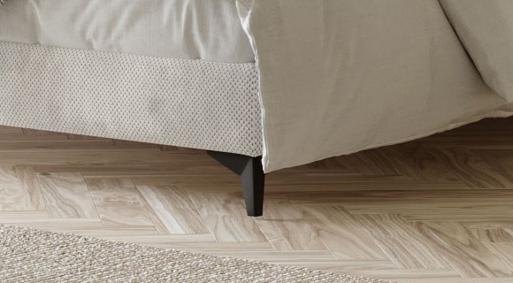 Detailaufnahme des unteren Bettrands eines Boxspringbetts: heller, beigefarbener Stoffbezug mit feiner, punktförmiger Struktur am Sockel, darüber eine locker fallende Bettdecke. Sichtbar ist ein dunkler, konisch zulaufender Bettfuß. Der Boden besteht aus hellem Holzparkett in Fischgrät-Optik; links unten schließt ein naturfarbener, grob gewebter Teppich an. Die Nahaufnahme betont Material, Verarbeitung und die Kombination von Textilien und Bodenbelägen