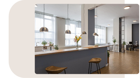 Moderner Pausenbereich mit einer langen Theke in Blau und Holz, dekoriert mit Blumenvasen. Über der Theke hängen mehrere Metallpendelleuchten. Zwei Barhocker stehen vor der Theke, und im Hintergrund sind weitere Sitzgelegenheiten und Pflanzen zu sehen. Große Fenster lassen viel natürliches Licht in den Raum und schaffen eine freundliche Atmosphäre