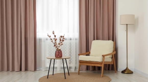 Sessel aus Holz mit hellem Polster neben Beistelltisch mit Vase vor pastellfarbenen Vorh&auml;ngen und Fenster.