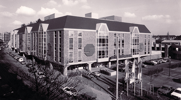 Das Bild zeigt ein gro&szlig;es Wohnkaufhaus mit markanter Architektur und geneigten Dachfl&auml;chen aus der Vogelperspektive. Die Fassade besteht aus einer Kombination aus Mauerwerk und gro&szlig;fl&auml;chigen Fensterfronten, die sich &uuml;ber mehrere Stockwerke erstrecken. Vor dem Geb&auml;ude sind Fahnenmasten, parkende Fahrzeuge sowie angrenzende Stra&szlig;en zu sehen. Die Aufnahme vermittelt einen &Uuml;berblick &uuml;ber das Geb&auml;ude und dessen st&auml;dtisches Umfeld.