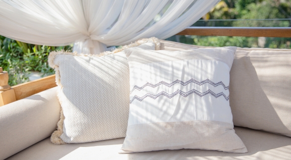 Beige Outdoor-Sofa mit zwei hellen Kissen, davor wei&szlig;er Vorhangstoff als Baldachin; Sonnenlicht wirft Streifen-Schatten auf die Polster.