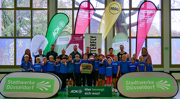 Das Bild zeigt eine gr&ouml;&szlig;ere Gruppe von Kindern und Erwachsenen, die gemeinsam in einer Sporthalle f&uuml;r ein Gruppenfoto posieren. Die Kinder tragen einheitliche blaue T-Shirts, w&auml;hrend hinter ihnen mehrere Banner und Fahnen verschiedener Partner und Organisationen aufgestellt sind. Im Vordergrund sind Werbebanden mit Logos zu sehen. Die Aufnahme vermittelt den Eindruck einer gemeinschaftlichen oder sportbezogenen Veranstaltung in einer Innenhalle.