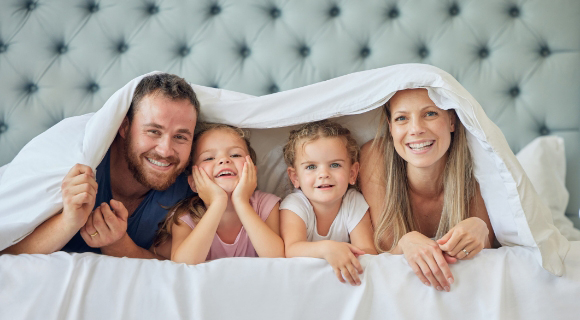 Lachende Familie liegt gemeinsam unter einer Decke auf einem großen Polsterbett mit gestepptem Kopfteil