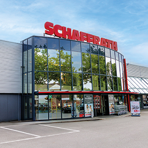 Das Bild zeigt den Eingangsbereich eines modernen Wohnkaufhauses mit einer gebogenen Glasfassade und dem Schriftzug &bdquo;Schaffrath&ldquo; oberhalb des Eingangs. Gro&szlig;e Fensterfl&auml;chen erm&ouml;glichen den Blick in den Innenraum und spiegeln B&auml;ume sowie den Himmel wider. Vor dem Geb&auml;ude befinden sich ein gepflasterter Vorplatz sowie Einkaufsk&ouml;rbe nahe dem Eingang. Die Aufnahme zeigt das Geb&auml;ude bei Tageslicht aus frontaler Perspektive.