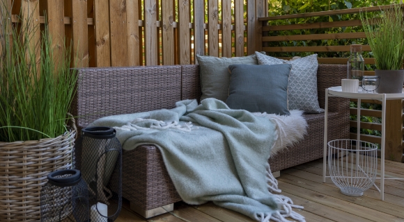 Ein gem&uuml;tlicher Balkon aus Holz mit einem geflochtenen Sofa auf dem t&uuml;rkise Kissen und eine Decke liegen, daneben steht ein runder Balkontisch mit Deko.