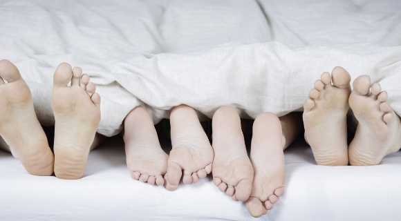 Zwei Erwachsene und zwei Kinder liegen unter einer weißen Decke, nur ihre nackten Füße ragen hervor.