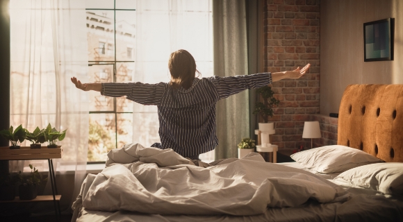 Frau streckt sich morgens im Bett vor einem großen Fenster – stimmungsvolles Schlafzimmer mit gepolstertem Kopfteil, Ziegelwand und natürlichem Lichteinfall