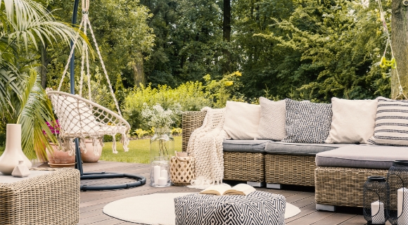 Rattan-Ecksofa mit H&auml;ngesessel und hellem, rundem Teppich auf einer Terrasse. 