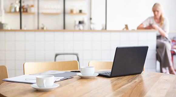 Helle Büroküche mit einem großen, runden Holztisch im Vordergrund, auf dem ein aufgeklappter Laptop, zwei weiße Kaffeetassen und ein Notizblock mit Kugelschreiber liegen. Im Hintergrund ist eine Küchenzeile mit weiß gefliester Front, Holzarbeitsplatte und offenen Regalen zu sehen. Auf den Regalen stehen Tassen, Gläser und weitere Küchenutensilien. Eine Frau mit blonden Haaren sitzt unscharf im Hintergrund auf einem Hocker und schaut auf ihr Smartphone. Die Szene vermittelt eine entspannte Arbeitsatmosphäre, in der sich Arbeit und Kaffeepause verbinden lassen.