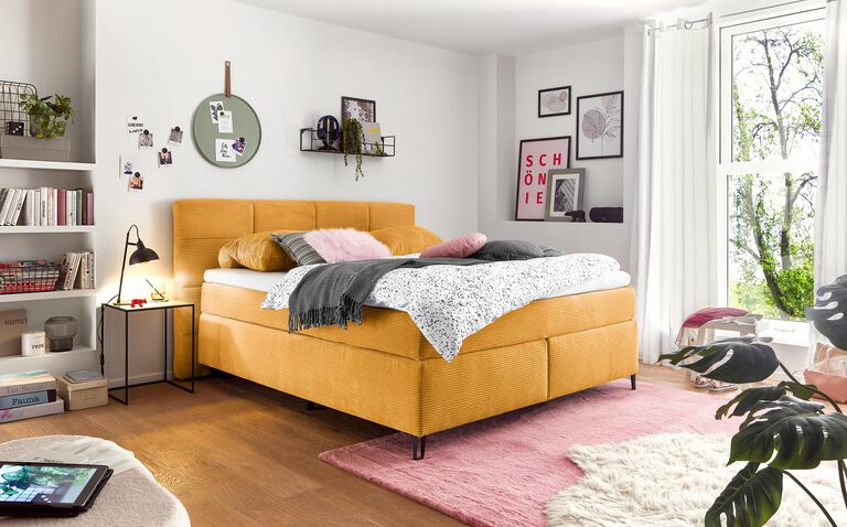 Schlafzimmer mit senfgelbem Bett, farbigen Deko-Details, Holzfußboden, weichen Teppichen in Rosa und Creme sowie großem Fenster.