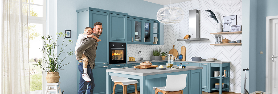 Ein Vater steht, sein Kind auf dem Rücken tragend, in einer hellblauen Kueche, welche im modernen Landhausstil gehalten ist