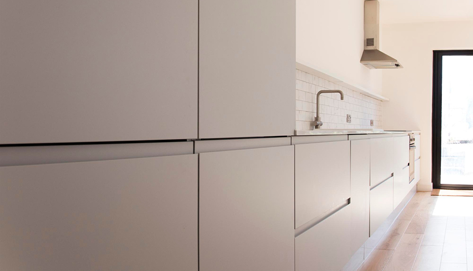 Minimalistische, helle Küche mit glatten, grifflosen Fronten in zartem Grau. Die lange Küchenzeile erstreckt sich entlang einer Wand und ist mit einer schlichten Arbeitsplatte sowie einem modernen Wasserhahn ausgestattet. Darüber verläuft ein weißer Fliesenspiegel mit rechteckigen Fliesen im Metro-Stil. Am Ende der Zeile befinden sich ein Einbauherd und eine Edelstahl-Dunstabzugshaube. Rechts im Bild fällt Tageslicht durch eine bodentiefe Glastür auf den hellen Holzboden und lässt den Raum offen und freundlich wirken. Die Gesamtwirkung ist klar, funktional und reduziert.
