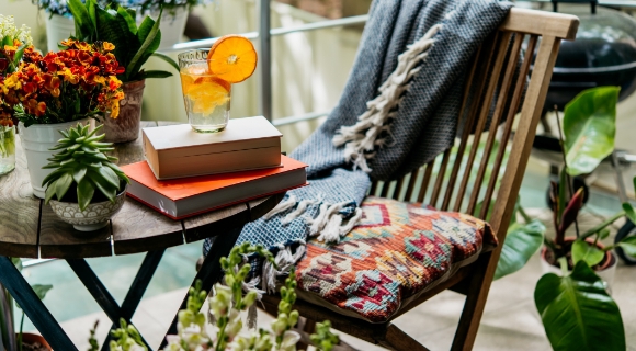 Kleiner Balkontisch mit B&uuml;chern und Glas mit Orangenscheibe neben Holzstuhl mit Decke, umgeben von Pflanzen