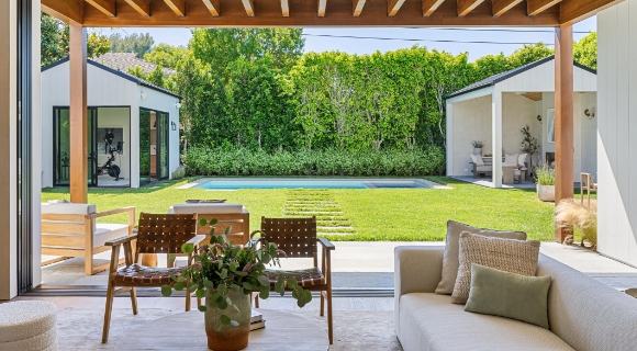 Blick aus dem Wohnbereich auf &uuml;berdachte Terrasse und Garten mit Rasenfl&auml;che, Pool und Sitzbereichen. 