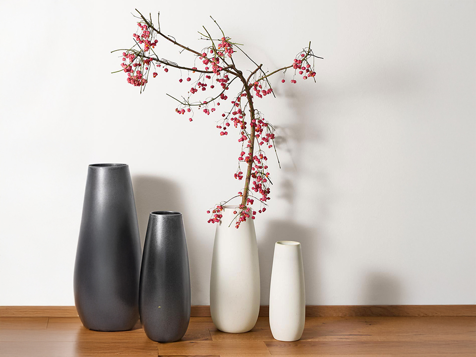 Vier hohe Keramikvasen in Schwarz und Weiß stehen auf einem Holzboden vor einer weißen Wand. Eine der weißen Vasen ist mit einem Zweig mit roten Beeren dekoriert.