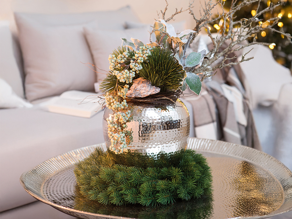 Auf einem dekorativen Tablett steht eine silberne Metallvase mit gehämmerter Oberfläche. Sie ist weihnachtlich mit Tannengrün, Beeren und Zweigen geschmückt und steht vor einem Sofa im Hintergrund.