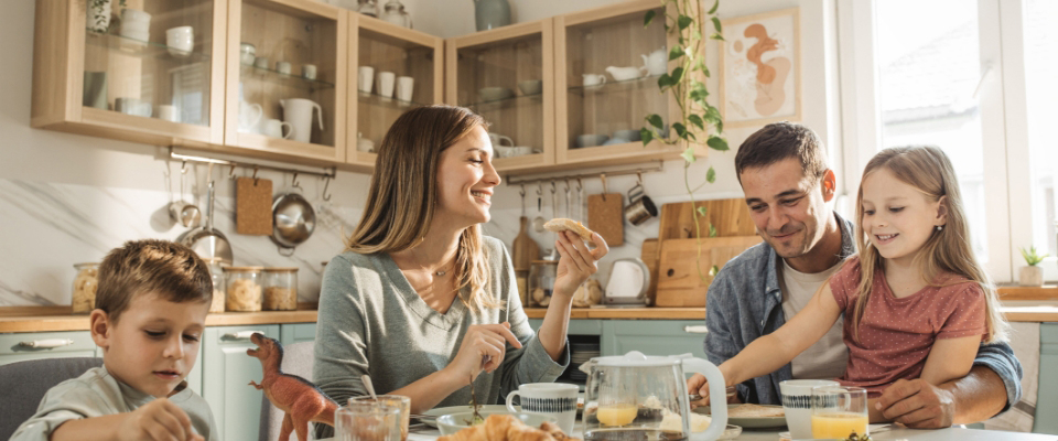 Eine vierköpfige Familie – bestehend aus Mutter, Vater, Sohn und Tochter – sitzen gemeinsam am Esstisch in der Küche und frühstücken.