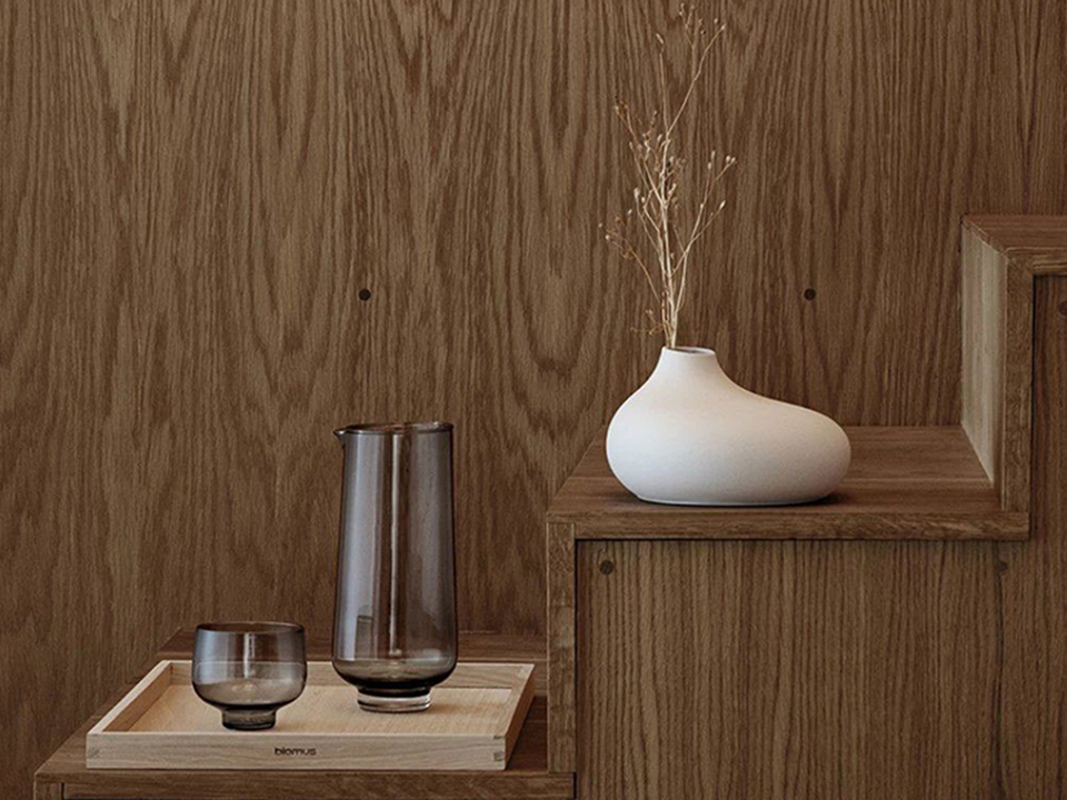 Eine weiße, bauchige Keramikvase mit Trockenblumen steht auf einer hölzernen Stufe. Daneben befindet sich ein Tablett mit einem Glas und einer Karaffe aus grauem Glas, passend zur minimalistischen Einrichtung.