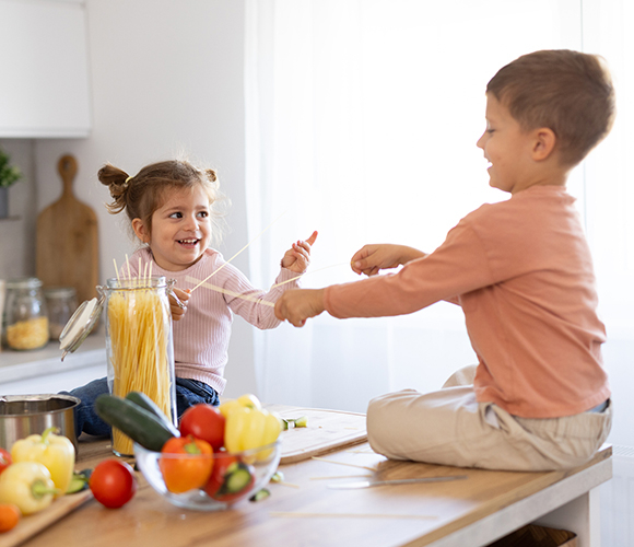Zwei Kinder spielen in der Küche mit rohen Spaghetti auf einer Kücheninsel voller Gemüse.