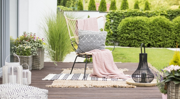 Sitzbereich auf einer Holzterrasse mit schwarzem Metallstuhl, rosa Decke, gemustertem Kissen und arrangierten Outdoor-Accessoires.