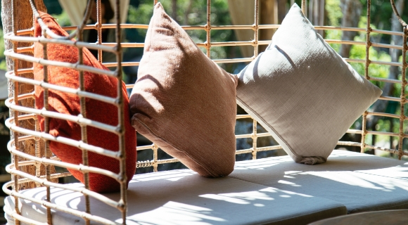 Geflochtener H&auml;ngesessel mit Sitzpolster und drei Kissen in Beige-, Braun- und Rott&ouml;nen.