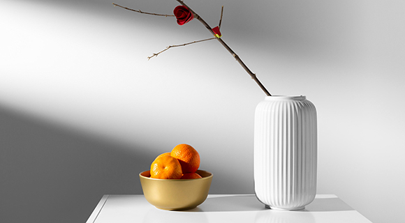 Auf einem weißen Tisch steht eine geriffelte weiße Vase mit einem einzelnen Zweig und roter Blüte. Daneben befindet sich eine goldene Schale mit mehreren Orangen, schlicht in Szene gesetzt.