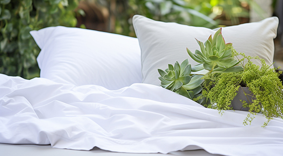 Bett mit weißer Sommerbettwäsche und zwei weißen Kissen im Grünen, dekoriert mit einem Topf aus Sukkulenten und filigranen Pflanzen