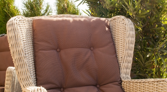 Nahaufnahme eines hellen Flechtsessels mit dickem braunen Sitzpolster; im Hintergrund gr&uuml;ne Str&auml;ucher im Sonnenlicht.