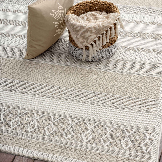 Beiger Outdoorteppich mit dezentem Streifen- und Strukturmuster auf einer Holzterrasse. Der Teppich liegt vor einer Sitzgruppe aus Holz und Seilgeflecht mit beigen Polstern und dekorativen Kissen. Im Hintergrund eine Natursteinmauer und ein gr&uuml;ner Rasenbereich.