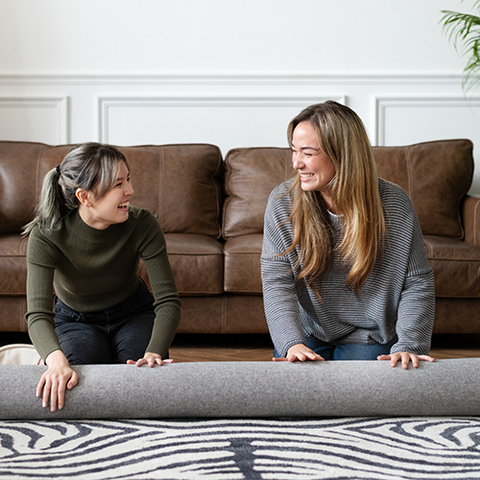 Zwei lächelnde Frauen rollen gemeinsam einen Teppich in einem modernen Wohnzimmer mit Ledersofa aus.
