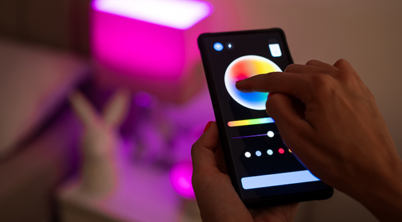 Hand bedient ein Smartphone, um das Licht einer farbwechselnden Lampe im Hintergrund zu steuern. 