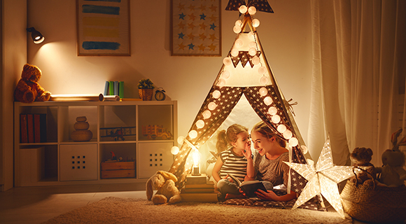 Mutter und Kind sitzen in einem sanft beleuchteten Kinderzimmer in einem Spielzelt mit Lichterketten und lesen ein Buch.