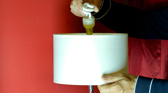 Hände setzen eine neue Glühbirne in eine Tischlampe mit weißem Schirm ein.