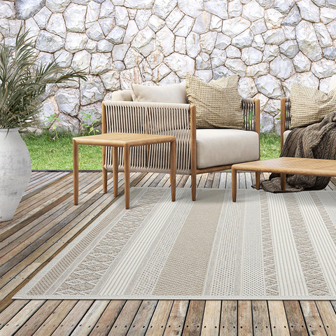 Beiger Outdoorteppich mit dezentem Streifen- und Strukturmuster auf einer Holzterrasse. Der Teppich liegt vor einer Sitzgruppe aus Holz und Seilgeflecht mit beigen Polstern und dekorativen Kissen. Im Hintergrund eine Natursteinmauer und ein grüner Rasenbereich.