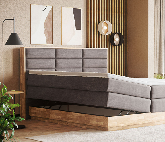 Modernes Boxspringbett in Mittelgrau mit gepolstertem Kopfteil in breiten, horizontalen Kassetten. Die Liegefläche ist elektrisch verstellt: im Rückenbereich deutlich angehoben, im Beinbereich leicht abgesenkt; obenauf liegt ein weißer Topper. Das Bett steht auf einem massiven Holzpodest, zwischen Box und Sockel ist die Verstellmechanik sichtbar. Links ein schlanker Beistelltisch mit schwarzer Stehleuchte, davor eine grüne Pflanze. Im Hintergrund eine Akzentwand aus vertikalen Holzlamellen, zwei runde Wandleuchten und abstrakte Bilder in Beige- und Sandtönen. Heller Teppichboden und warme Naturfarben erzeugen eine ruhige, moderne Schlafzimmeratmosphäre