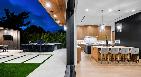 Moderne offene Wohnküche mit Holz- und Schwarzelementen, angrenzender Terrasse und Outdoor-Küche bei Abendbeleuchtung.