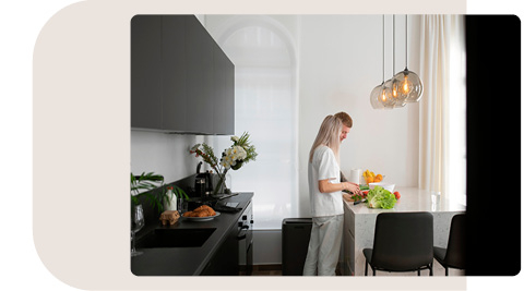Paar in moderner Küche mit dunklen Schränken und heller Arbeitsfläche. Die Frau bereitet frisches Gemüse vor, während der Mann neben ihr steht. Die Küche ist mit Blumen dekoriert, und über der Arbeitsfläche hängen drei stilvolle Pendelleuchten. Große Fenster lassen natürliches Licht in den Raum
