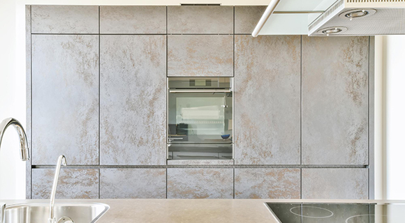 Moderne Einbauküche mit grifflosen Fronten in realistischer Betonoptik. Die gesamte Wand ist mit großflächigen, nahtlos aneinandergereihten Schrankelementen verkleidet, mittig ist ein Einbaubackofen mit Glastür integriert. Im Vordergrund befindet sich eine Kücheninsel mit glatter Arbeitsfläche, integriertem Edelstahl-Spülbecken und einem modernen Induktionskochfeld. Darüber ist ein Teil der Edelstahl-Dunstabzugshaube sichtbar. Die klare Linienführung, das minimalistische Design und die kühle Betonoptik verleihen der Küche einen puristischen, urbanen Charakter.